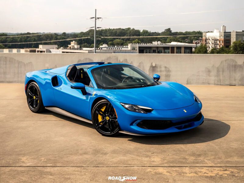 A blue Ferrari convertible sports car, showcasing one of the iconic Ferrari colors, is parked on a rooftop with city buildings and trees in the background.