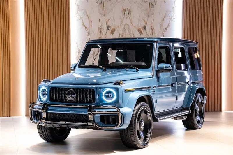 SUV Mercedes-Benz G-Class yang mewah diparkir di dalam ruangan, diterangi oleh pencahayaan lembut. Kendaraan itu, sempurna untuk dijual, menawarkan desain kotak dan gril depan yang menonjol.