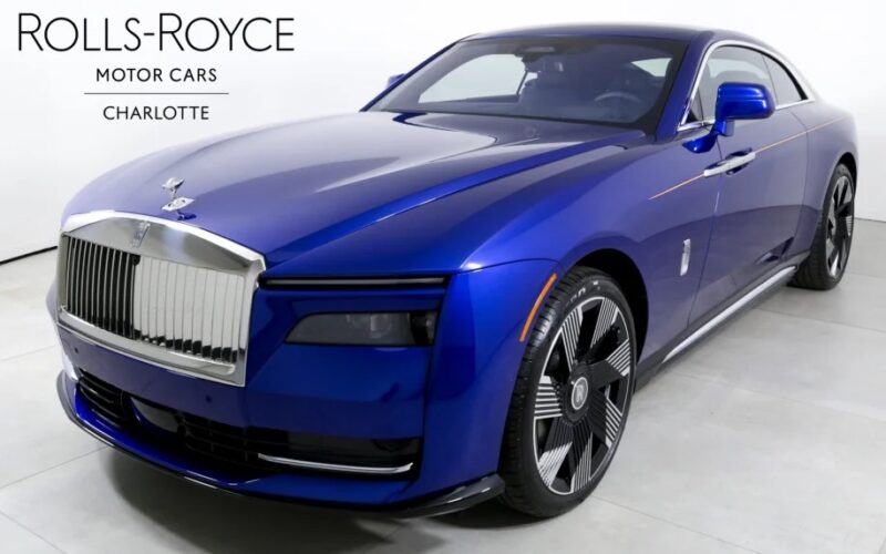 A blue Rolls-Royce Spectre coupe, an electric ultra-luxe super coupe, is displayed indoors; text in the corner reads "Rolls-Royce Motor Cars Charlotte.