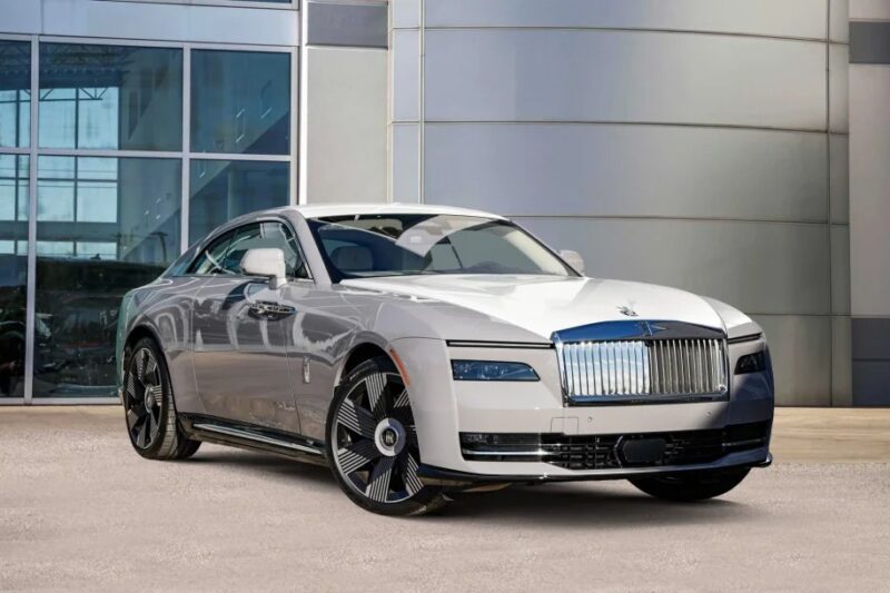 A silver Rolls-Royce Spectre, an ultra-luxe electric super coupe, is parked in front of a modern glass building.