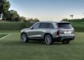 A sleek silver Cadillac SUV, a luxury EV model, is parked on a grassy field surrounded by trees and palm trees under a clear sky.