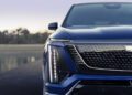 Close-up of the front of a blue Cadillac Vistiq SUV, showcasing the left headlight, grille, and part of the hood against a blurred natural background, highlighting its luxury EV design.