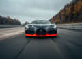 A sleek black Bugatti sports car with red accents, possibly the famed W16 Mistral, speeds down a highway surrounded by trees on a cloudy day, hinting at its potential to set a new world record.