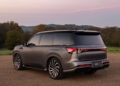 An Infiniti SUV with a matte gray finish is parked on a dirt road, trees and hills silhouetted against a pinkish sky. Its presence exudes the essence of tailgating luxury, inviting thoughts of adventure and tranquility intertwined.