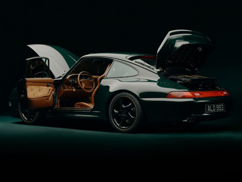 A dark green Porsche 993 Turbo sports car with its doors, front hood, and rear engine compartment open, showcasing a tan interior and black-rimmed wheels.