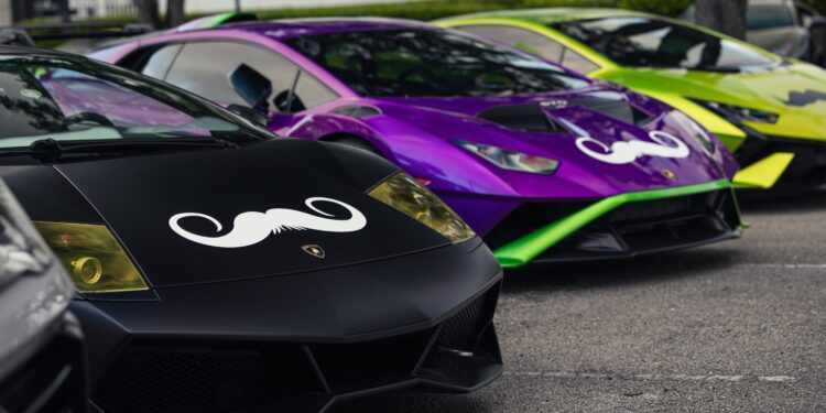 Three sleek sports cars, including a striking Lamborghini, are parked in a row, each proudly sporting a large mustache decal on the hood in support of Movember. The vibrant colors—black, purple, and green—make a bold statement for Men's Health awareness.