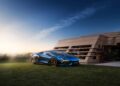 A sleek blue Lamborghini sports car, a new model debut, is parked on a grassy lawn next to a modern house at sunset.