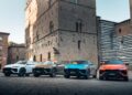 Four SUVs in white, gold, blue, and orange, including a Lamborghini new model debut, parked in front of historic stone buildings on a cobblestone street.