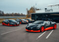 Four sleek black and orange W16 Mistral Bugatti sports cars are parked in front of a building adorned with "Bugatti" signage. Trees and a road form the backdrop, hinting at the brand's world-record ambitions.