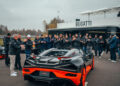 A crowd applauds as a black and orange Bugatti sports car, possibly the stunning W16 Mistral, is parked in front of a building with a Bugatti sign—a scene of excitement perhaps worthy of a world record.