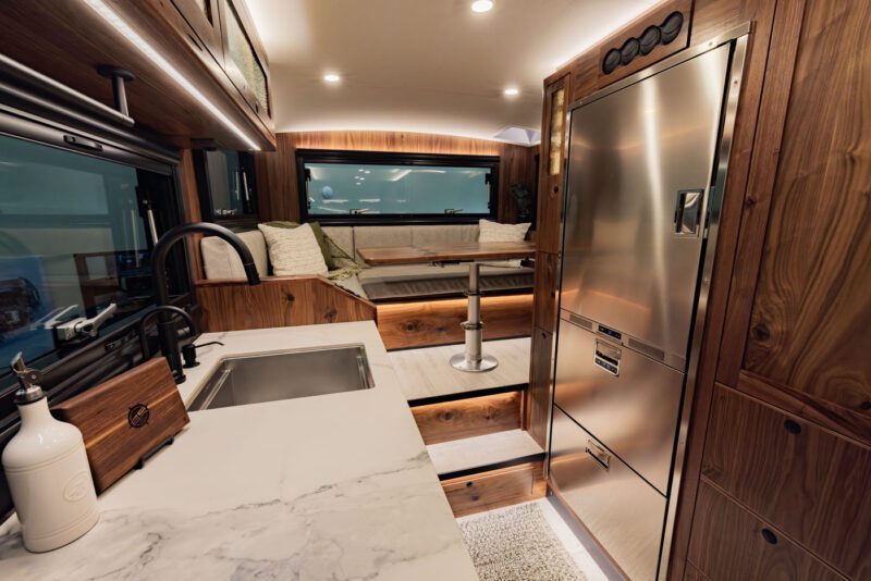 The interior of this EarthRoamer LTx expedition vehicle boasts a modern design with a marble countertop, wooden cabinetry, a sink, and a cozy seating area by large windows. This is the ultimate redefined space for those who seek adventure without sacrificing comfort.