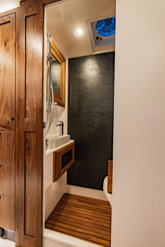 Compact bathroom with wooden accents, perfect for an EarthRoamer expedition vehicle. It features a small sink, mirror, and shower area. The dark, textured walls complement the floor's wooden slat design, creating a cozy yet adventurous vibe.