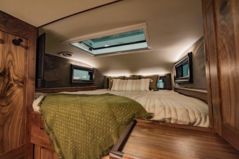 The cozy bedroom interior of the EarthRoamer LTx camper van boasts a snug bed adorned with a green blanket, complemented by elegant wood paneling and a skylight above, perfect for any expedition vehicle adventure.