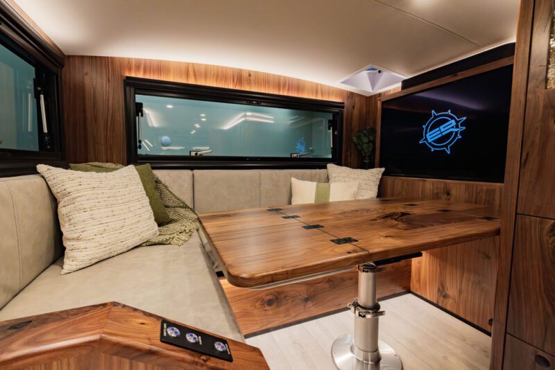 Interior of a modern Expedition Vehicle with wooden accents, featuring a cozy seating area with cushions, a foldable table, and a TV screen on the right wall. This ultimate design ensures comfort and style for every adventure.