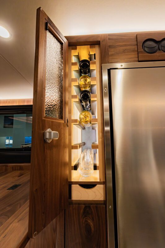 A wooden cabinet with a door ajar reveals a vertical wine rack, an ultimate touch of luxury, holding four bottles and a glass positioned in front of it—a nod to the refined comforts found in an EarthRoamer expedition vehicle.