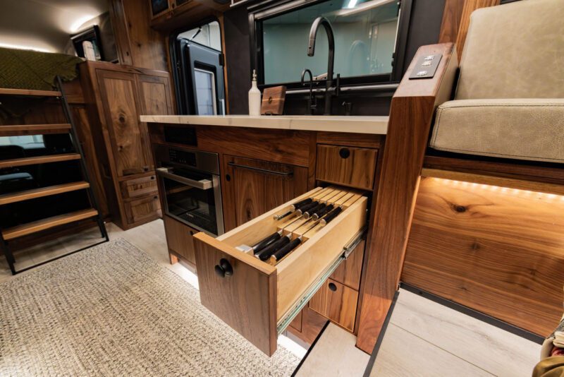 Modern kitchen with wooden cabinetry, a drawer open revealing organized knives, and a sink with black fixtures, perfectly suited for an EarthRoamer LTx. This expedition vehicle’s interior combines elegance with functionality for the ultimate adventure-ready lifestyle.