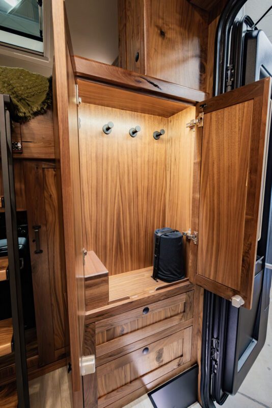 The wooden cabinet interior of the EarthRoamer LTx expedition vehicle features three hooks on the back panel. A small black cylindrical object rests on the bottom shelf, while the open cabinet door reveals its sturdy structure, perfect for storing essentials during your adventurous travels.