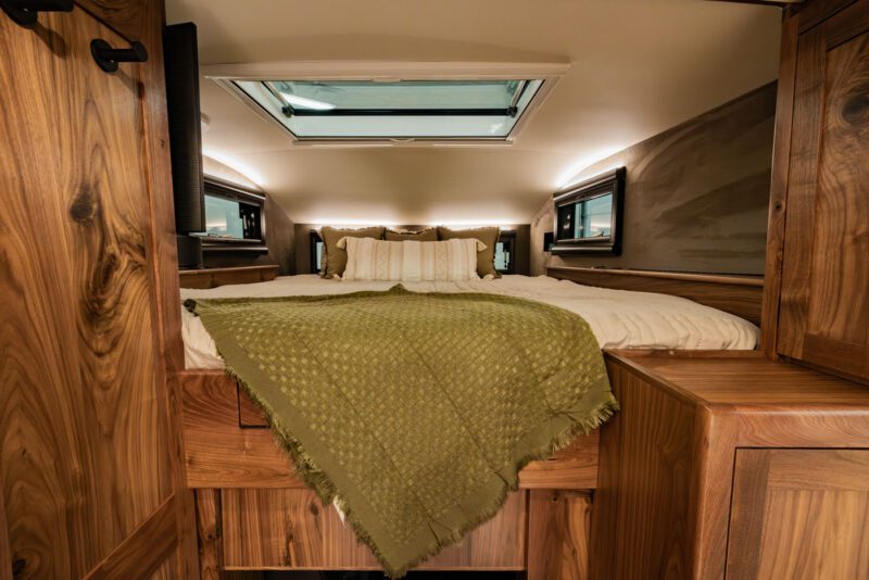 Cozy wooden bedroom inside an EarthRoamer expedition vehicle, featuring a skylight above a neatly made bed. An olive green blanket is draped at the foot of the bed, creating a serene and adventurous retreat.