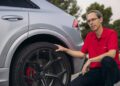 A man in a red shirt, possibly a chassis developer, kneels beside a silver Audi RS Q8 Performance. He's focused on inspecting a wheel with a distinctive black rim and red brake caliper. Majestic trees stand tall in the background, creating an intriguing scene that hints at Spanish influence.