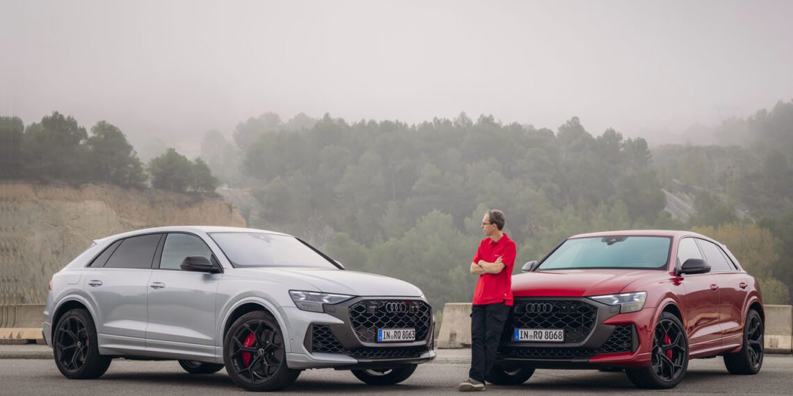 A person, possibly a chassis developer, stands next to two parked Audi SUVs—an elegant gray and a striking red—on a foggy Spanish road. Each vehicle exudes the performance prowess of the Audi RS Q8.