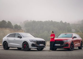 A person, possibly a chassis developer, stands next to two parked Audi SUVs—an elegant gray and a striking red—on a foggy Spanish road. Each vehicle exudes the performance prowess of the Audi RS Q8.