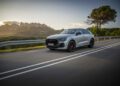 A sleek silver Audi SUV cruises along a scenic road, its RS Q8 Performance on full display against a backdrop of majestic mountains and towering trees, all under a clear blue sky.