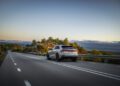 A white Audi RS Q8 Performance glides along a scenic, empty road in Spain under a clear sky, surrounded by lush greenery and distant mountains.