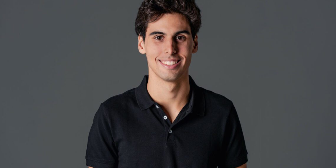 A person with short dark hair, reminiscent of F1 Driver Gabriel Bortoleto, stands against a gray background in a black polo shirt, smiling confidently.