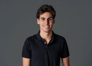 A person with short dark hair, reminiscent of F1 Driver Gabriel Bortoleto, stands against a gray background in a black polo shirt, smiling confidently.