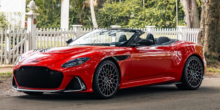 A red sports car from The Collection is parked on a shaded street, flanked by a white picket fence and trees in the background, reminiscent of classic Aston Martins.
