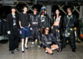 A group of six people poses indoors wearing stylish, streetwear-inspired outfits from the new collection, including oversized jackets, sporty pants, and sunglasses. They stand in a line against a modern industrial backdrop reminiscent of A$AP Rocky's edgy aesthetic.