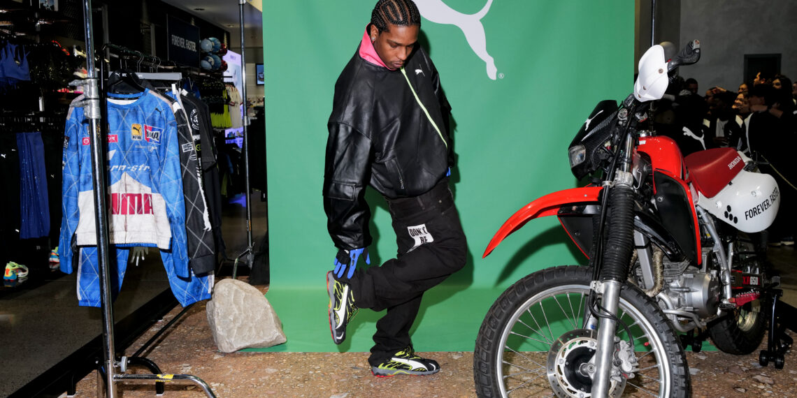 A person in a black jacket poses in front of a green Puma Motorsport backdrop, alongside A$AP Rocky's new collection display, next to a red and white motorcycle and a clothing rack.