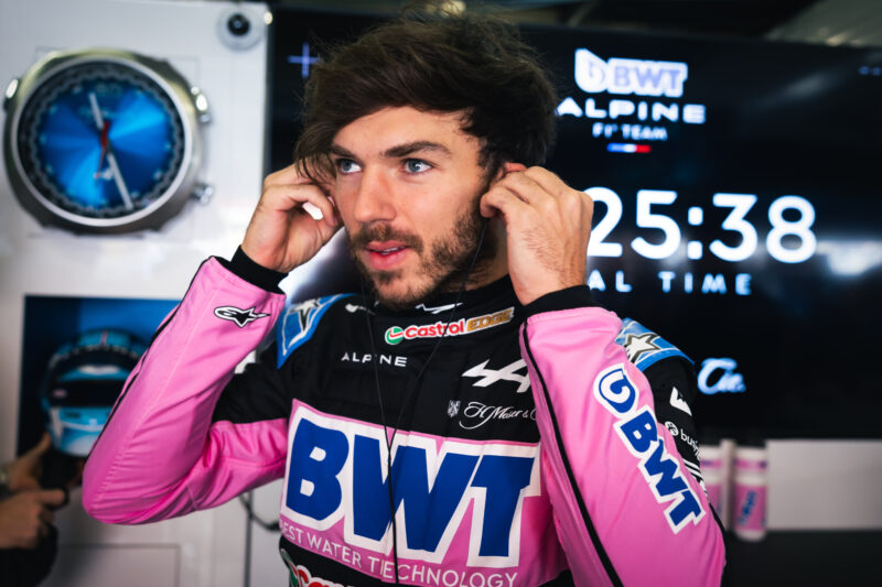 Race car driver in a black and pink BWT Alpine suit adjusts his earbuds on the starting grid, with a digital clock displaying 25:38, ready to kick off the 2025 F1 season.