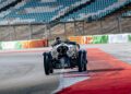 A vintage Bentley Speed Six with the number 22 races on a track, surrounded by empty stands and colorful barriers, reminiscent of its glory days.