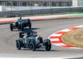 Two vintage cars racing on a track, the foreground featuring a classic Bentley displaying the Union Jack and number plate J 1926, reminiscent of the iconic Speed Six from its Continuation Series.