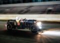 A vintage black Bentley Speed Six racing car with headlamps on, driven by a helmeted driver, speeds along the track, echoing the classic elegance of its Continuation Series design.