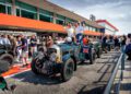 A classic Bentley Speed Six adorned with a Union Jack is parked at an event. Two people stand in the car, holding bottles, as spectators admire the Continuation Series alongside other vintage cars in the background.