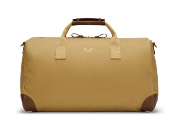 Tan duffel bag with brown leather handles and corner accents, featuring a logo on the front, epitomizes luxury accessories. Elevate your style upgrade with this sophisticated piece—now available for sale.