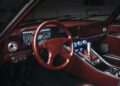 Vintage car interior featuring a red leather steering wheel, classic analog gauges, and a metal gear shifter surrounded by illuminated controls, reminiscent of driving a manual supercar with the iconic TWR V12 engine.