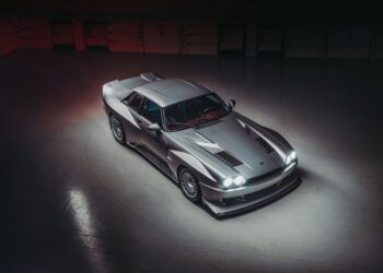 Aerial view of a metallic silver sports car with pop-up headlights, resembling a Supercat. Parked in a dimly lit indoor space, its V12 engine hints at unmatched power, while the sleek design and aerodynamic features are highlighted.