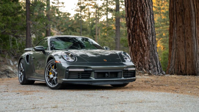 A dark gray sports car parked on a forest road, surrounded by tall trees.