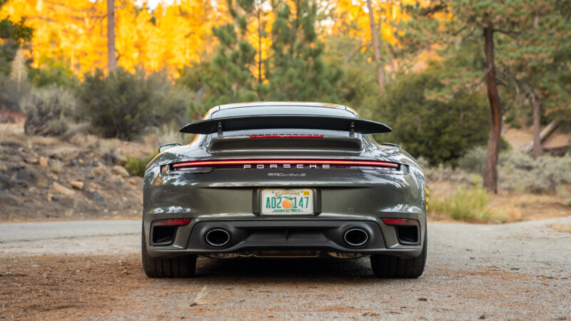 Rear view of a gray Porsche sports car parked on a rural road, featuring dual exhausts and a spoiler, with autumn trees in the background.