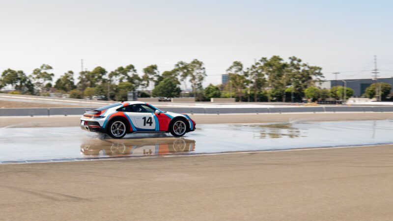 A sports car with number 14 on the side skids on a wet track during a driving test. Trees and a building are visible in the background.