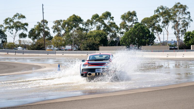 A red car drives through a water-covered track, creating a large splash. Trees and a concrete barrier are visible in the background.