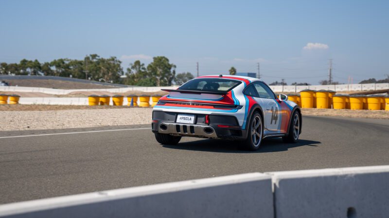 A sports car with colorful racing stripes is driving on a race track. Yellow barriers and trees are in the background.