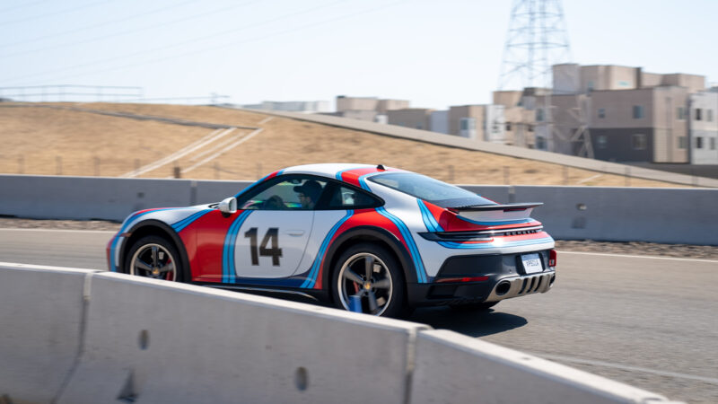 A sports car with number 14 and colorful stripes drives on a racetrack with barriers. Buildings and a hill are in the background.