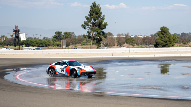 Car number 14 in colorful livery skids on a wet track during a driving event, with a figure standing by the track holding a sign in the background.