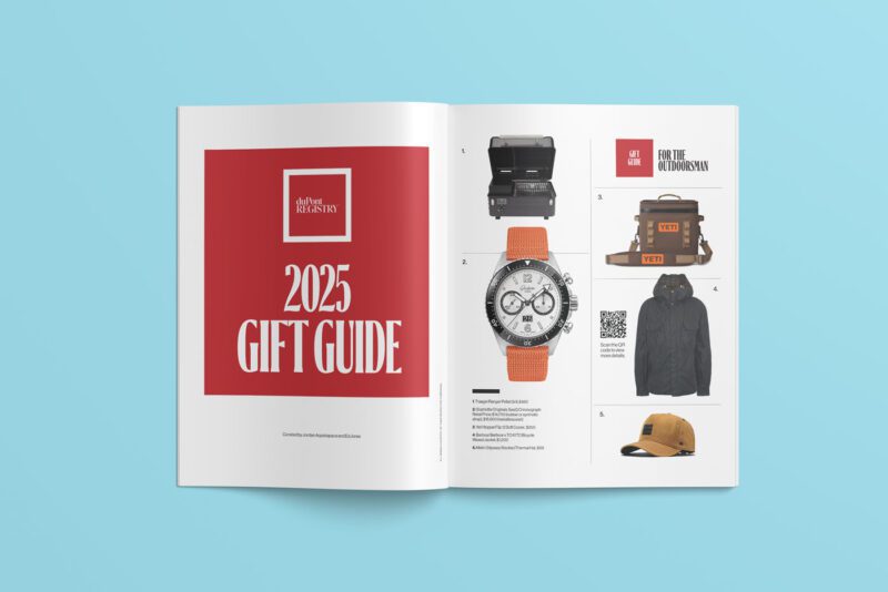 Open magazine revealing duPont REGISTRY's "2025 Gift Guide Edition" with a striking red cover. The right page showcases an array of gift items like a watch, jacket, camera, and hat against a light blue backdrop. Perfect for your December 2024 shopping inspirations.