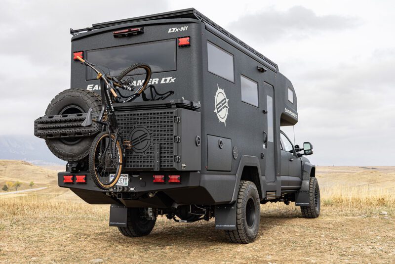 An ultimate black expedition vehicle, resembling an EarthRoamer, stands parked in an open field. With a bike and spare tire mounted on the back, it looks ready to conquer any terrain under the vast, cloudy sky.