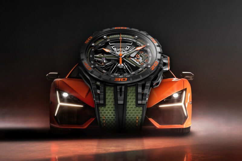 An ultimate chronograph with an intricate design is integrated into the front of an orange sports car, highlighting speed and technology, set against a dramatic dark background.